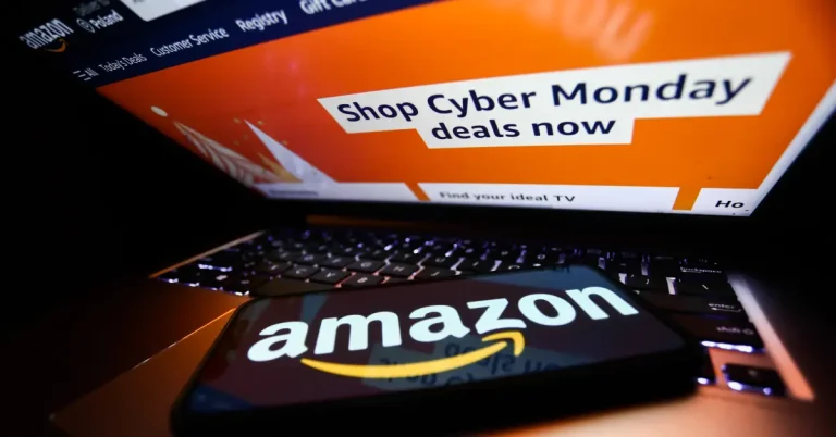 A partially open laptop displaying Amazon's website with the words shop Cyber Monday deals. A phone displaying the Amazon logo rests on the laptop's keyboard.