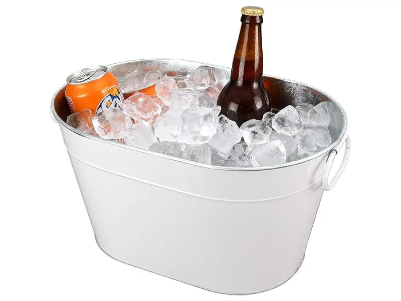 White oval metal beverage tub filled with ice cubes, chilling one brown glass bottle and one orange soda can, with metal handles on the sides, on a plain white background.